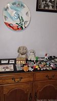 Photo of assorted keychains, small video games, anime figures, and stuffed animals laid out on wooden furniture against white wall with abstract wall plate.