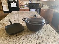 Photo of two cookware pieces on a granite kitchen counter: a square griddle pan with long handle and a large round Dutch oven with lid and side handles.