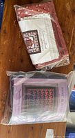 Two separate quilting kits in sealed plastic bags. One has red and cream fabrics with a 'Crossing Threads' pattern, the other has black and purple fabrics with a 'Tessellating Hounds Tooth' pattern.