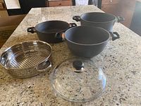 Photo of three cookware pieces and one steamer insert on granite countertop: two Dutch ovens, one glass lid, one stainless steel vegetable steamer.