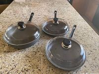 Three SCANPAN fry pans of different sizes (8", 10.25", 12.5") on granite counter, each with a glass lid and black Bakelite handle.