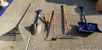Photo showing all six tools laid out on pavement including metal leaf rake, shovel, hedge trimmers, garden hoe, metal garden rake, and blue plastic snow shovel.