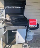 Weber Genesis grill open showing grill grate, vegetable basket, cookbook, and open cabinets underneath.