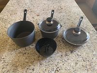 Three black SCANPAN sauce pans with black handles; two have glass lids and one is without a lid. One black perforated veggie steamer insert is placed in front of the pans on a granite countertop.