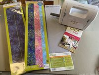 Photo showing the GO! Baby fabric cutter, two acrylic templates in packaging, a 6 x 24 cutting mat, and user manual all displayed on a table