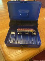 Open blue metal cash box showing five metal coin compartments and removable tray inside lid.