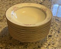 Stack of eight cream-colored Noritake Magnificence pattern fine china soup bowls with floral detailing and gold trim on a granite countertop.