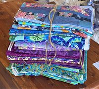 Photo of stacked bundles of quilting cotton fabrics tied with twine, showing predominantly blue, purple, and green colors with various floral and abstract patterns