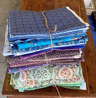 Three bundles of neatly folded quilting cotton fabric cut lengths tied with twine, displayed on a wooden table showing blue, purple, green, and mixed prints.