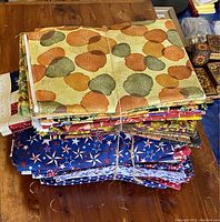 First image of the two fabric bundles laid on a wooden table. Top bundle primarily autumn colors with repeating pumpkin and leaf patterns, bottom bundle patriotic star print fabrics in blue, red, and white.
