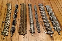 Seven vintage bracelets laid out side by side on wood surface showing various metal link and design styles including filigree, mesh, and enamel accents.