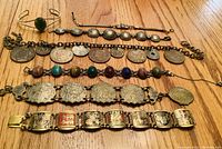 Photo showing 7 vintage bracelets laid out on a wooden surface, displaying various designs including coats of arms, old coins, scarabs, an oval jade piece, and Shakespeare scenes.