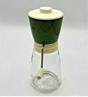 Side view of the vintage nut grinder showing the green metal grinding chamber, beige lid, crank handle, and clear glass container base.