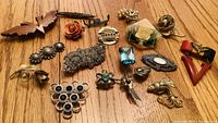 Top-down photo of 17 vintage style pins arranged on wooden surface. Pins feature various shapes including flowers, birds, geometric patterns, and butterflies. Colors predominantly metallic with some colored accents.