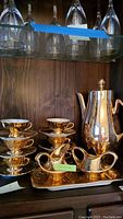 Full view of tea set displayed on wooden shelf, showing the teapot, stacked teacups with saucers, sugar bowl, creamer on a tray.