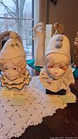 Two Cybis clown bust sculptures Funny Face and Valentine with Holly displayed on a lace tablecloth near window.