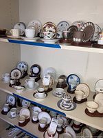 Multiple shelves displaying 15 assorted demitasse cups and matching small plates in various designs and brands.