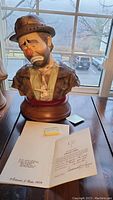 Photo of the handcrafted porcelain bust sculpture of sad clown wearing a hat with Certificate of Authenticity and artist's business card shown in front on table.