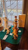 Photo of five figurines on a green cloth on a wooden table near window, includes Goebel angel, clown, and maiden figures