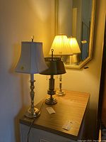 Three brass table lamps on wood topped cabinet, showing different shade styles and lamp base designs.