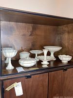 Full view of 8 Lenox milk glass platters, candy dishes, bowls on wooden shelf.