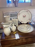 Photo showing various Lenox ceramic items including pitchers, vase, platters, collectors plate on stand, and small pourers grouped on wooden surface in front of window with air conditioner.