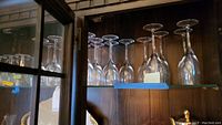 Sixteen upside-down stemmed glasses lined up on a shelf view, showing gold rims and slender stems.