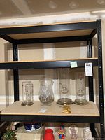 View of three cylindrical glass oil lamps with brass bases and one clear glass vase on shelving unit.