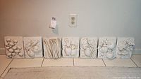 Seven white cast wall plaques with 3D fruit and vegetable designs set against a wall on tile floor, showing the full lot.