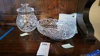 Two pieces of Waterford crystal on a wooden surface, bowl on the right and jar with lid on the left, certificate of authenticity behind bowl.