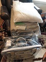 Stack of bedding items including pillows with striped pillowcases on top, beige quilted bedspread underneath, and a black and white plaid bedding set in a clear plastic carrying case at the bottom.