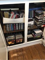 Cabinet shelf filled with multiple CDs, a box containing CDs, and stacks of audio tapes and boxed CDs.