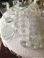 Top view of various crystal stemware glasses, tall clear glass vases, and a round embossed serving plate on white tablecloth.