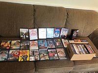 Multiple DVDs and a cardboard box filled with DVDs and CDs arranged on a brown couch including 'Once', 'The Full Monty', 'Vera', 'Midsummer Night's Dream', and more.