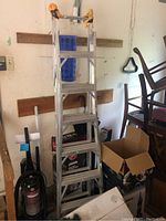 Full view of the aluminum extension ladder leaning against a wall with some surrounding objects such as vacuum cleaner, boxes and chairs.