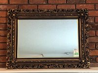 Framed wall mirror hanging on brick wall, front view showing the decorative frame and clear reflective surface.