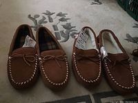Two pairs of brown moccasin slippers side by side on carpet showing whipstitch detail and bows.