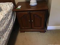 Photo of one wooden night stand with two cabinet doors, brown finish, showing top and front, positioned next to bed.