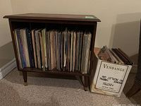 Wooden mid-century style cabinet filled with vinyl records plus a cardboard box with additional records beside it.