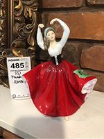 Porcelain figurine of a lady in a red skirt and white top with arms raised, tagged and placed against a warm brick background.