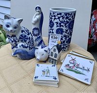 Photo showing the tall blue and white Delft style vase, three figurines (cat, pig, woman), small house figurine, and stacks of coasters on a beige tablecloth.