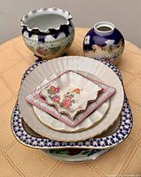 Stack of assorted plates and small dishes on a tablecloth. Includes floral and pink Wedgwood plate and small square pink floral dish, white bowl with blue and gold detailing, and blue and gold planter and vase behind them.