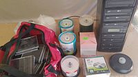 Wide view of LG CD/DVD duplicator tower, red bag filled with jewel cases, and multiple spindles of blank discs on table