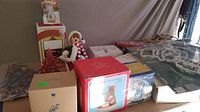 Wide view of Christmas decorations consisting of multiple boxed figurines, lighted ornaments, a fabric elf doll, and hanging lights arranged on a table.