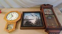 Three clocks side by side on a table: an octagonal Ingraham wooden clock, a square Daniel Dakota 3D wildlife clock, and a tall wooden Westminster chime clock with glass front and pendulum.
