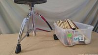 Photo of adjustable drum throne with tripod chrome legs and rubber feet, black padded seat beside plastic bin with drumsticks
