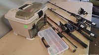 Photo showing five fishing rods with attached reels laid out on a table with two plastic fishing tackle boxes beside them.