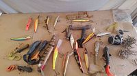 Overall view of fishing tackle laid out showing various muskie lures, soft plastic baits, fishing reel, and hooks.
