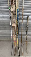 Photo displaying 11 hockey sticks with various brands and types leaning against a corrugated metal backdrop.