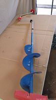 Full view of manual ice auger on a table showing blue spiral bit and red handle.